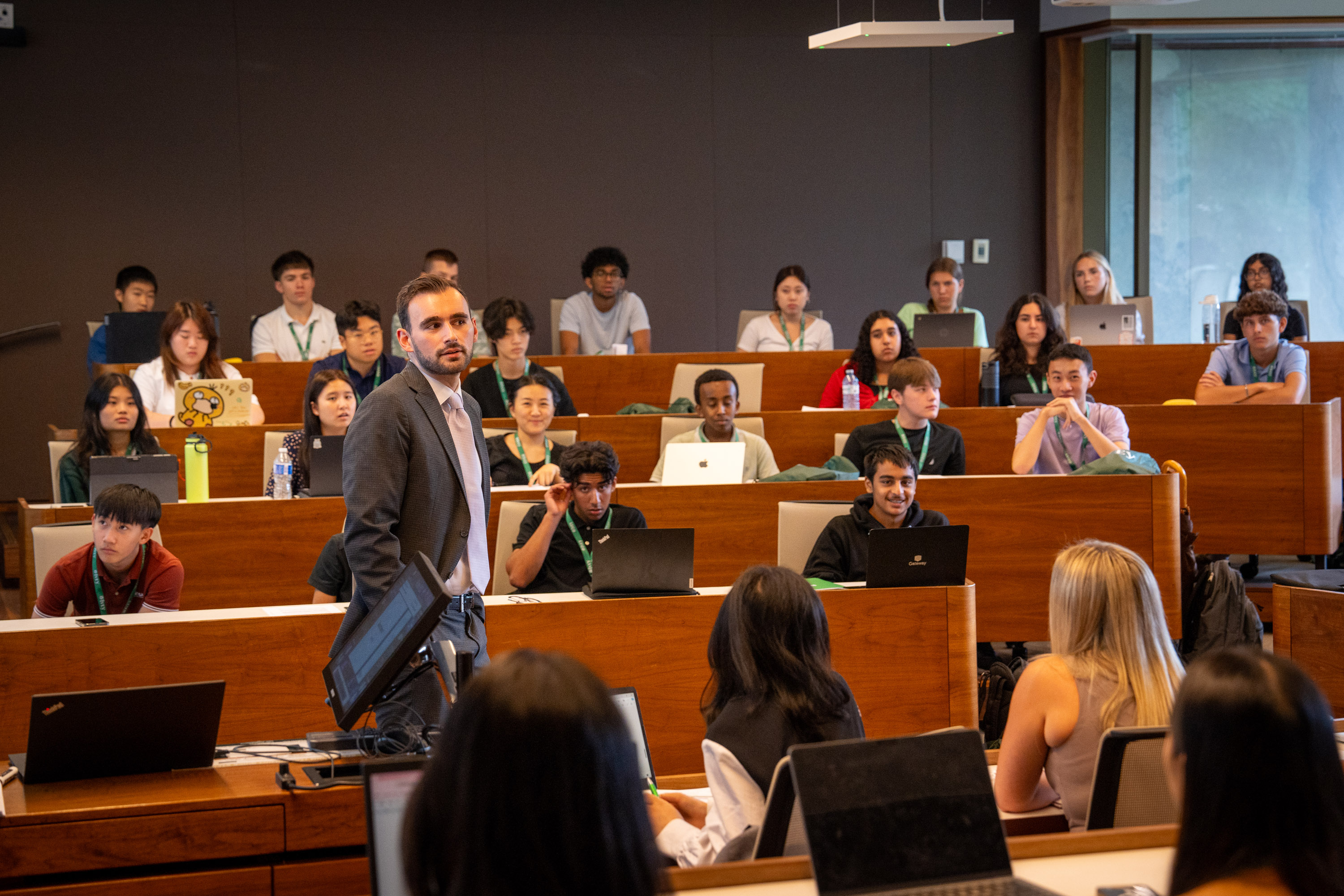 A tiered classroom full of high school students with laptops on the desks in front of them.  There is a male professor in the centre of the room, leading the discussion.