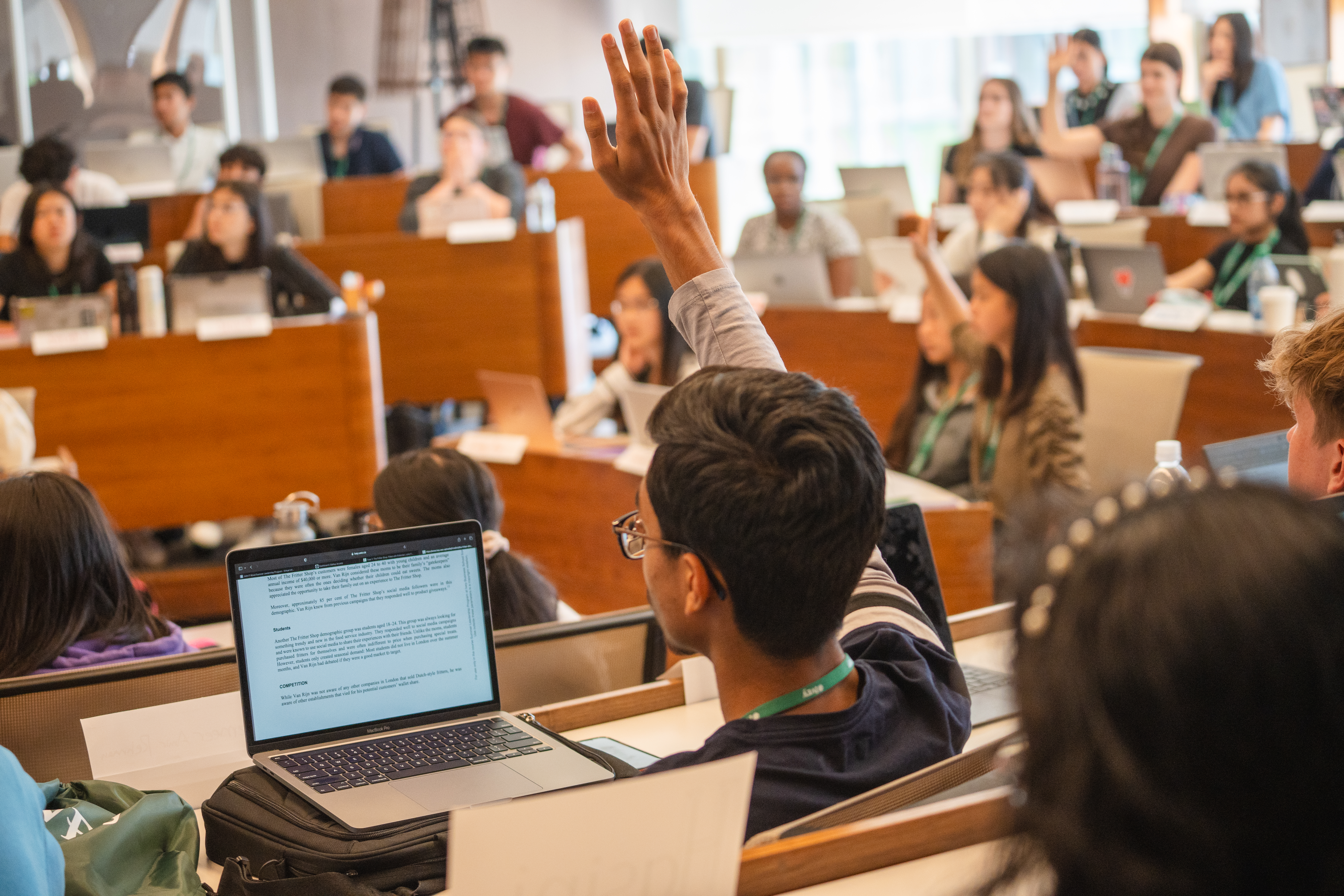 A tiered classroom full of high school students with  their hands raised eager to participate in the class discussion